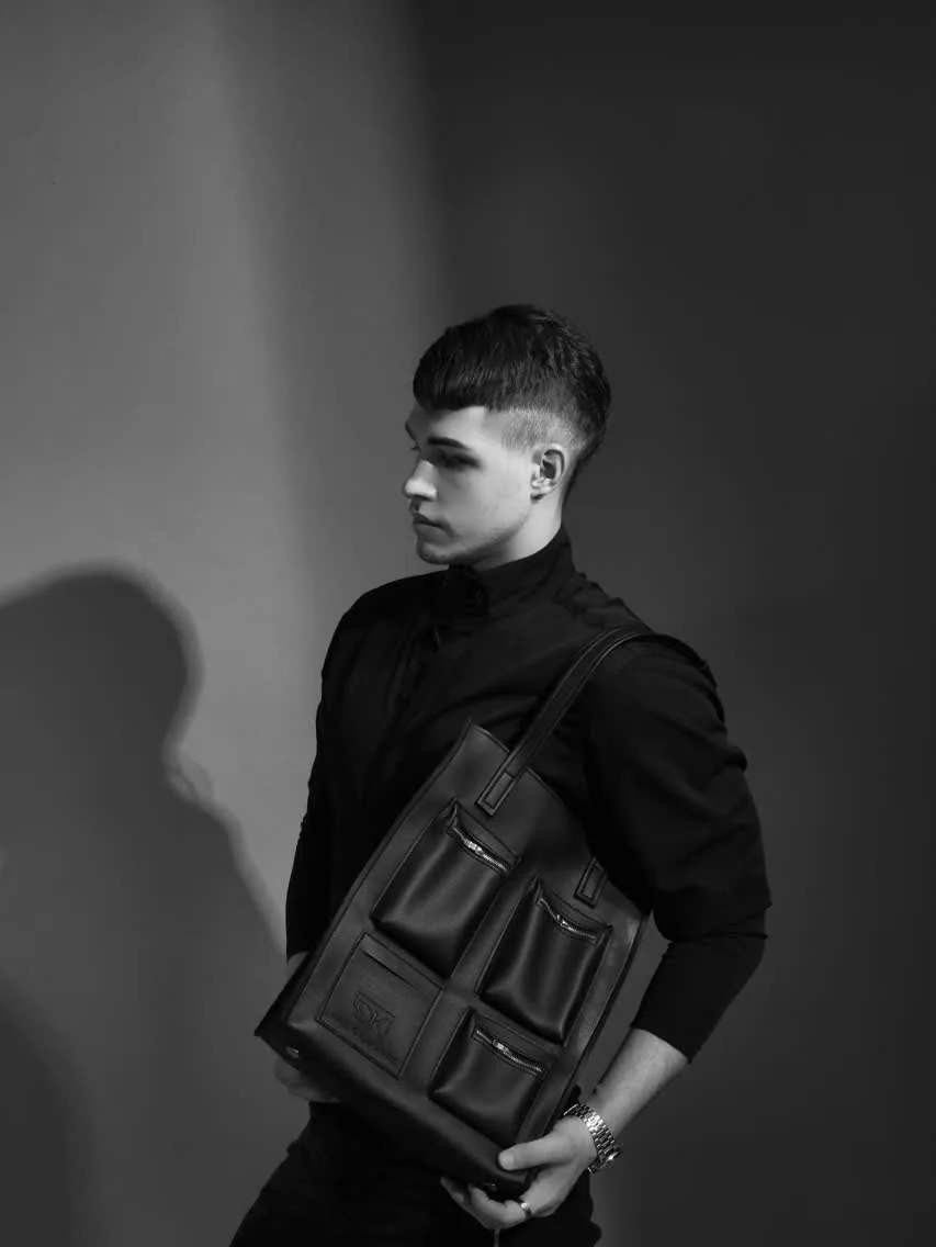 Black-and-white studio image of a model in profile carrying the DK 01 Moscow tote on the shoulder, his shadow on the wall behind.